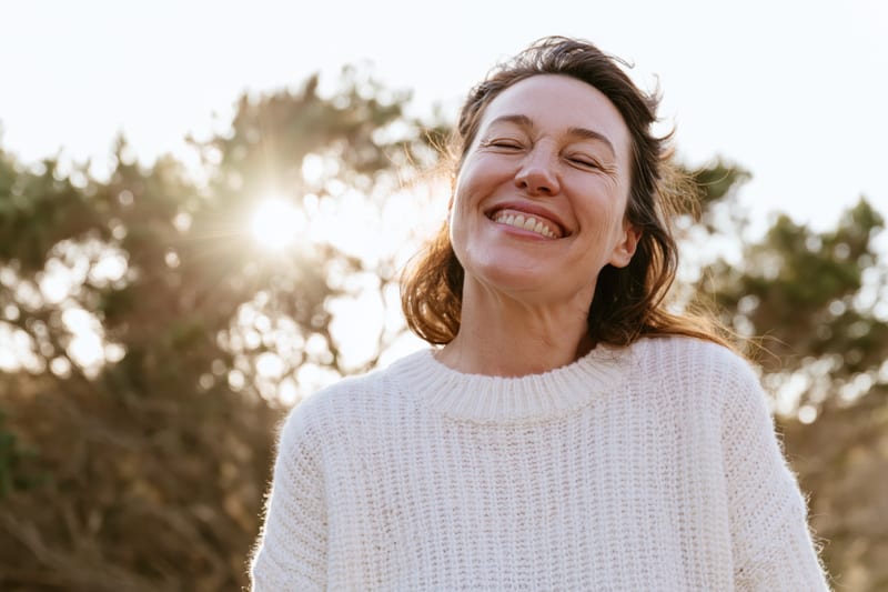 A smiling woman in a white knit sweater with her eyes closed, the sun shining through a tree behind her
